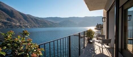 A balcony with a view of a lake and mountains, a table with chairs, and a potted plant.