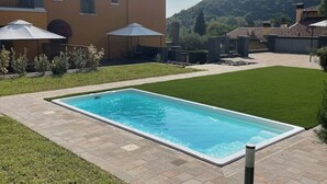 A rectangular swimming pool with a tiled deck, surrounded by a well-maintained lawn and a building with a tiled roof. - Villa Valle Franciacorta - Green Apartment with Pool and Garden (Rodengo-Saiano)