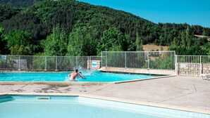 Une piscine avec une personne dans l’eau, entourée d’une clôture métallique et avec une colline boisée en arrière-plan.