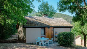 A small, single-story building with a thatched roof, surrounded by trees. There is a small table with chairs outside, and a colorful umbrella nearby. - Cottage for 8 People (La Canourgue)