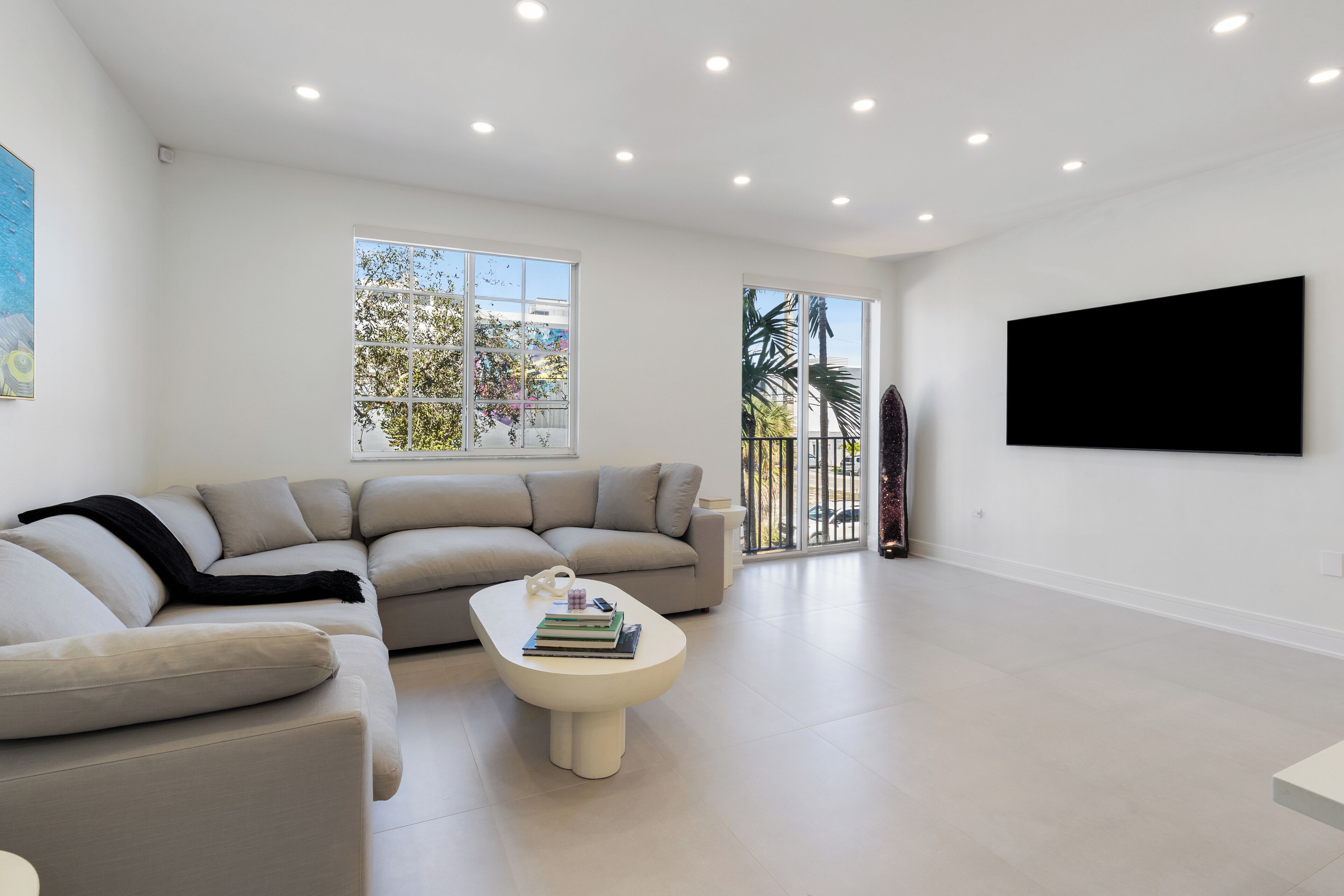 Una sala de estar moderna con un sofá color beige, una mesa de centro blanca con libros, y un ventanal con vistas a palmeras.