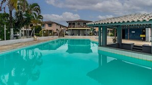 A swimming pool with a tiled deck, a covered seating area, and a building with a balcony in the background. - Apto com Varanda e Piscinas em Porto Seguro (Porto Seguro)