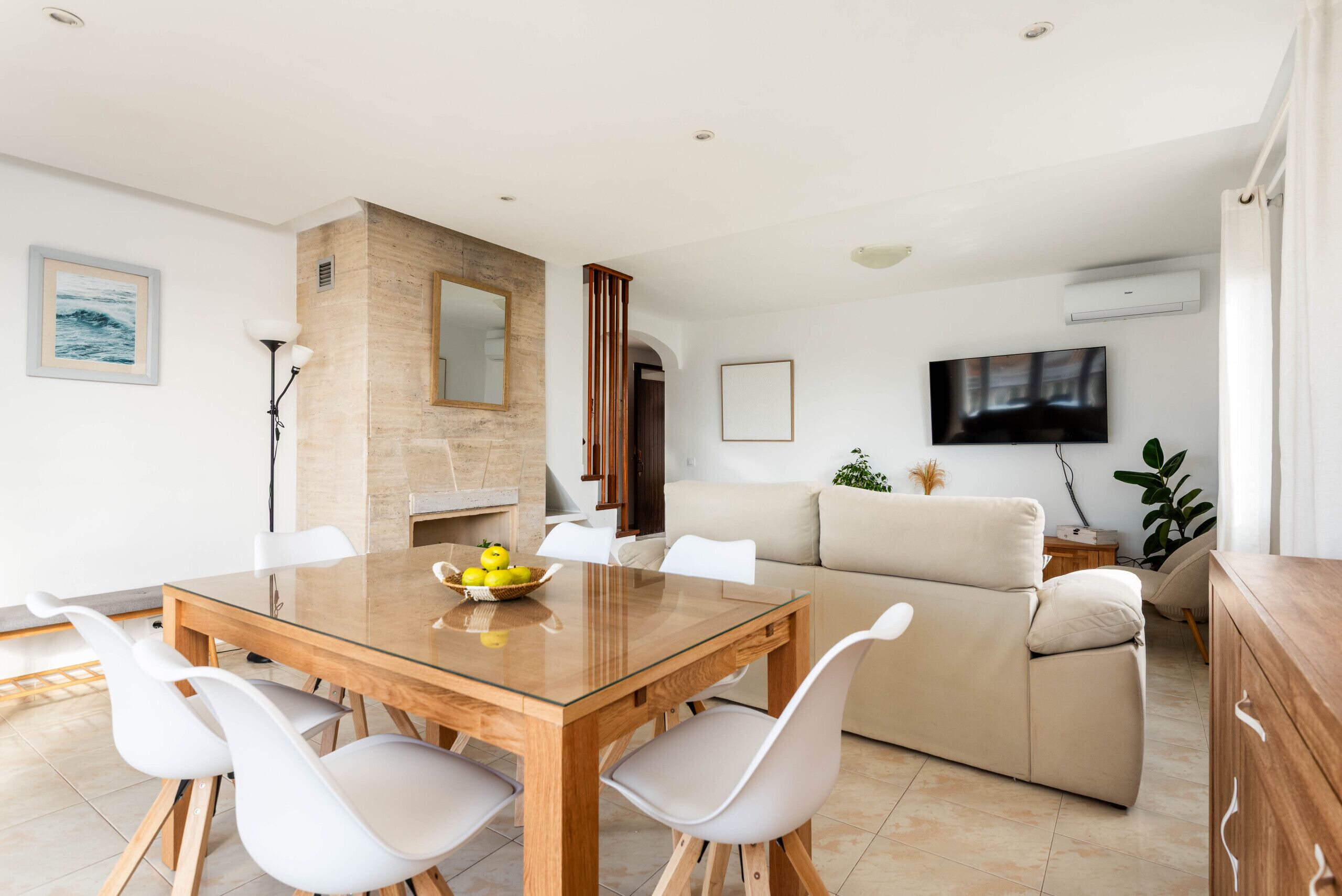 A modern living room with a wooden dining table, white chairs, a beige sofa, and a flat-screen TV mounted on the wall.
