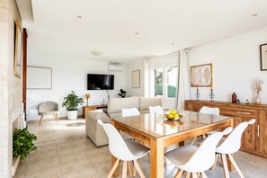 Ein modernes Wohnzimmer mit einem Esstisch mit Glasplatte, weißen Stühlen, einem beigen Sofa und einem flachbildfernseher an der Wand.