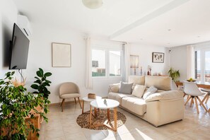 Ein modernes Wohnzimmer mit einer Couch, einem Couchtisch und einem an der Wand befestigten Fernseher. Es gibt Pflanzen, ein Fenster mit Vorhängen und einen Essbereich mit Stühlen.