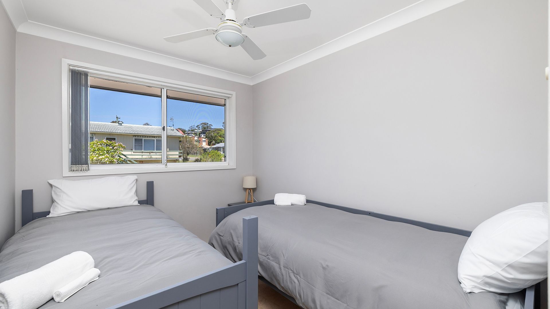 Un dormitorio con dos camas, un ventilador de techo, una ventana con vistas a casas y una mesita de noche con lámpara.