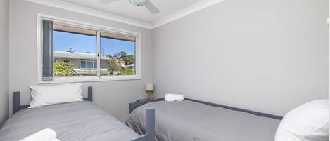 Un dormitorio con dos camas, un ventilador de techo, una ventana con vistas a casas y una mesita de noche con lámpara.