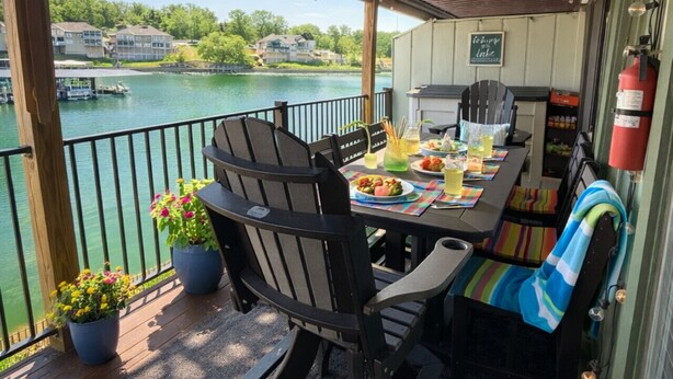 Un área de comedor al aire libre techada con una mesa preparada para dos, sillas, un ventilador de techo y vistas a un lago con barcos.