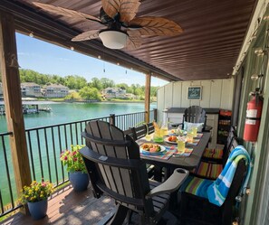 Un área de comedor al aire libre techada con una mesa preparada para dos, sillas, un ventilador de techo y vistas a un lago con barcos.