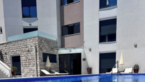 A swimming pool with a clear blue water surface, a glass of iced coffee on the poolside, and a building with multiple windows and balconies in the background.