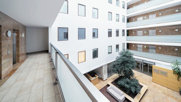 A modern building with multiple floors, a central courtyard with a seating area and a tree, and a reception desk. - RentPlanet - Kazimierza Wielkiego (Poznań)