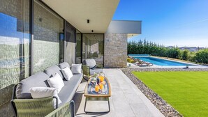 Una zona esterna moderna con divano, poltrone e un tavolo con bevande. In secondo piano, si vede una piscina.