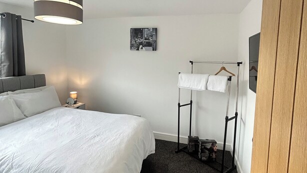 A hotel room with a large bed, a bedside table with a lamp, a hanging clothes rack, and a door. - cozy serene flat (Dudley)