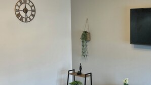 A modern living room with a ceiling light fixture, a wall clock, a small plant, a black TV stand, and a black sofa. - cozy serene flat (Dudley)