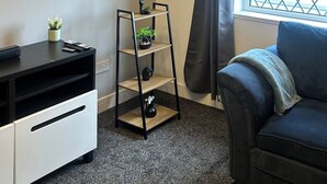 A modern living room with a black sofa, a small wooden shelf with plants, a black TV stand, and a window with curtains. - cozy serene flat (Dudley)