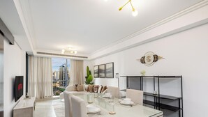 Ein modernes Wohnzimmer mit Essbereich, großen Fenstern und Blick auf die Stadt, einem Flachbildfernseher und einer dekorativen Uhr.
