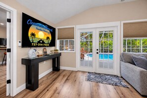 A living room with a pool view, a TV displaying a beach-themed image, and a wooden console table. - 3 min walk to beach/Heated Pool/Games/Kids area (Clearwater Beach)