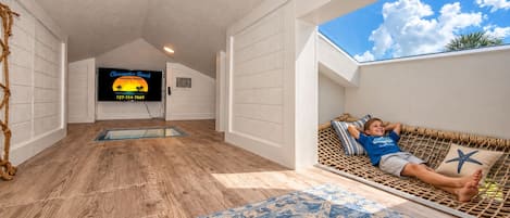 Una habitación con piso de madera, un televisor en la pared, un niño en una hamaca y una alfombra estampada.
