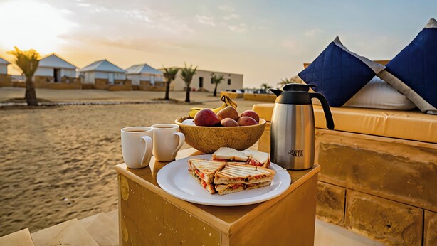Une table en bois dressée pour le petit-déjeuner, avec une assiette de sandwichs, un bol de fruits, une tasse de café et un thermos.