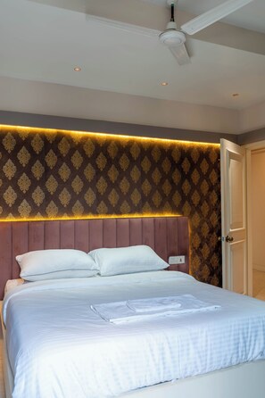 A neatly made bed with white linens and pillows, a headboard with a patterned design, and a ceiling fan. - BODHI BAARI (Kolkata)