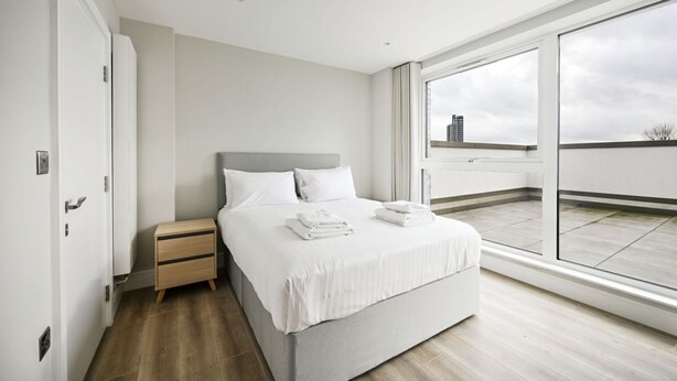A modern bedroom with a large window, a bed, a nightstand, and a wooden floor. - Argyl | Modern 2 Bed Apt. with Balcony & Elevator (Greater London)