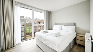 A modern hotel room with a large bed, a bedside table, and a view of the city. - Argyl | Modern 2 Bed Apt. with Balcony & Elevator (Greater London)