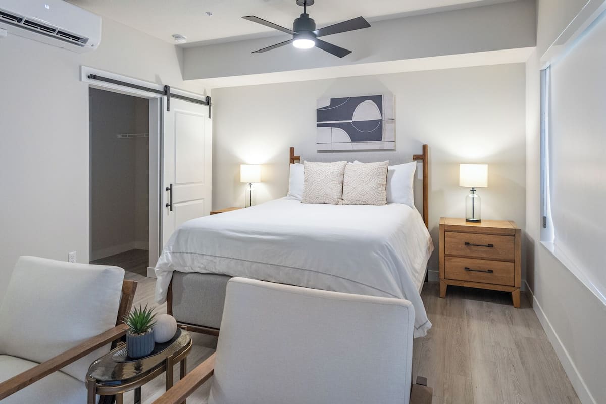 Ein Schlafzimmer mit einem Bett, Nachttischen, einem Sessel, einem Deckenventilator und einem an der Wand hängenden Kunstwerk.