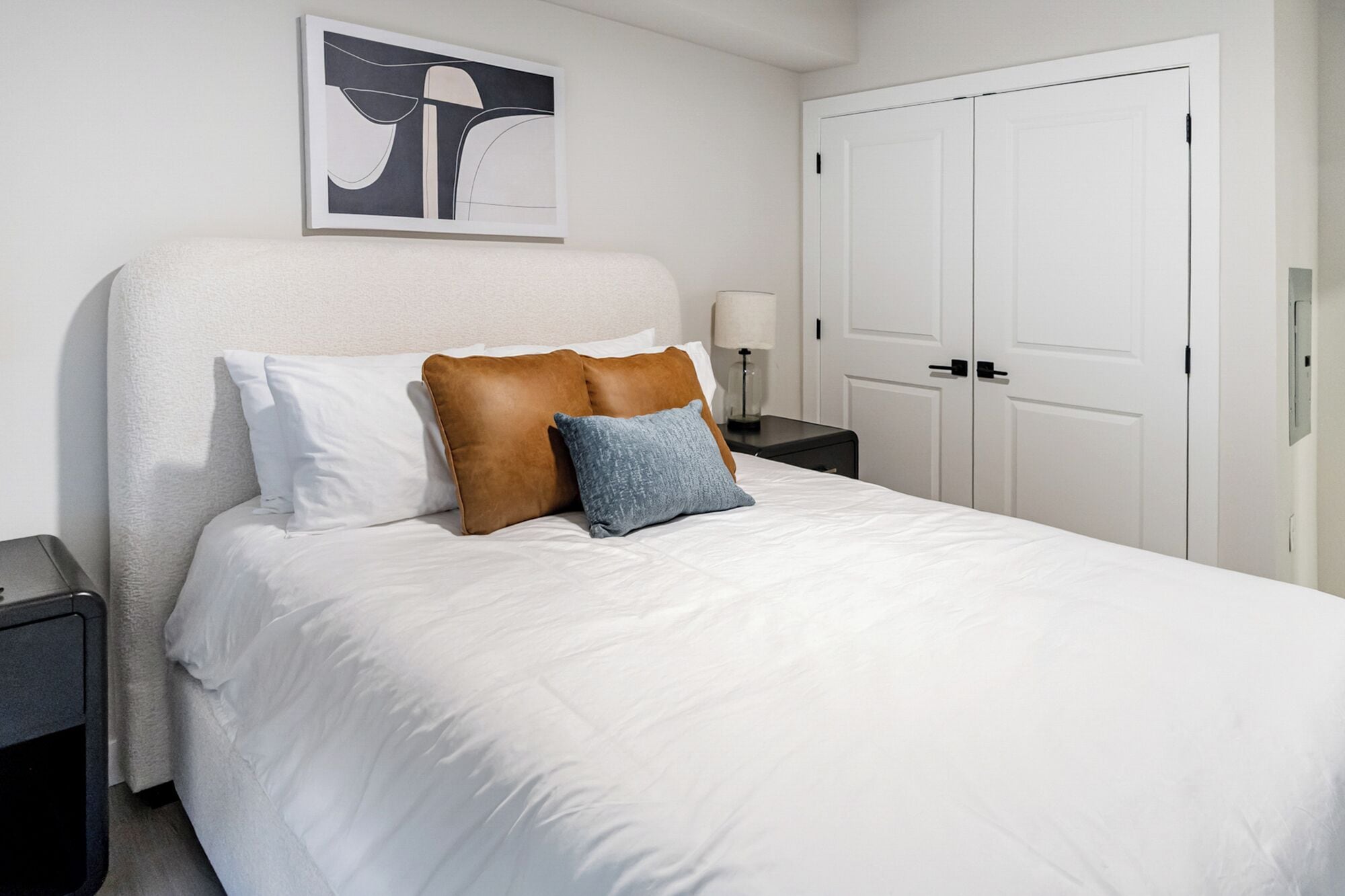 Una cama bien hecha con ropa blanca, una almohada marrón y azul, una lámpara de noche y una obra de arte abstracta enmarcada en la pared.