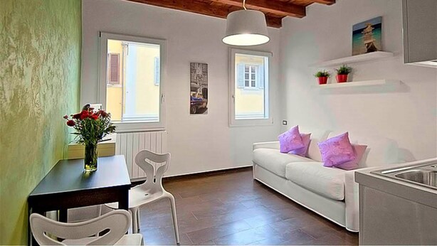 Une pièce avec un plafond en bois, des murs blancs, un canapé blanc agrémenté de coussins violets, une table à manger avec des chaises et un vase de fleurs.