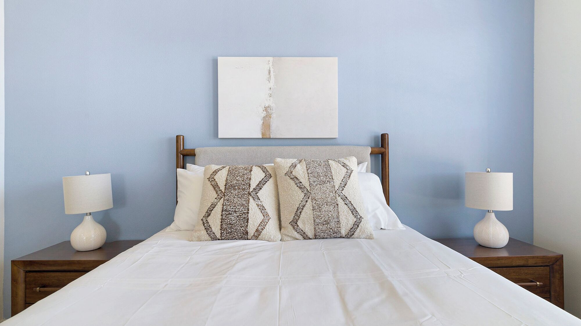 Una cama bien hecha con ropa blanca y dos almohadas estampadas, flanqueada por dos mesitas de noche con lámparas, frente a una pared azul claro.