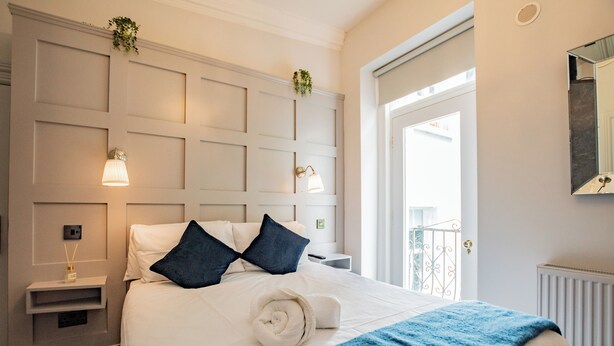 A hotel room with a neatly made bed, a bedside table with a lamp, and a view of the outside balcony. - Howth Harbour Cosy Retreat (Howth)