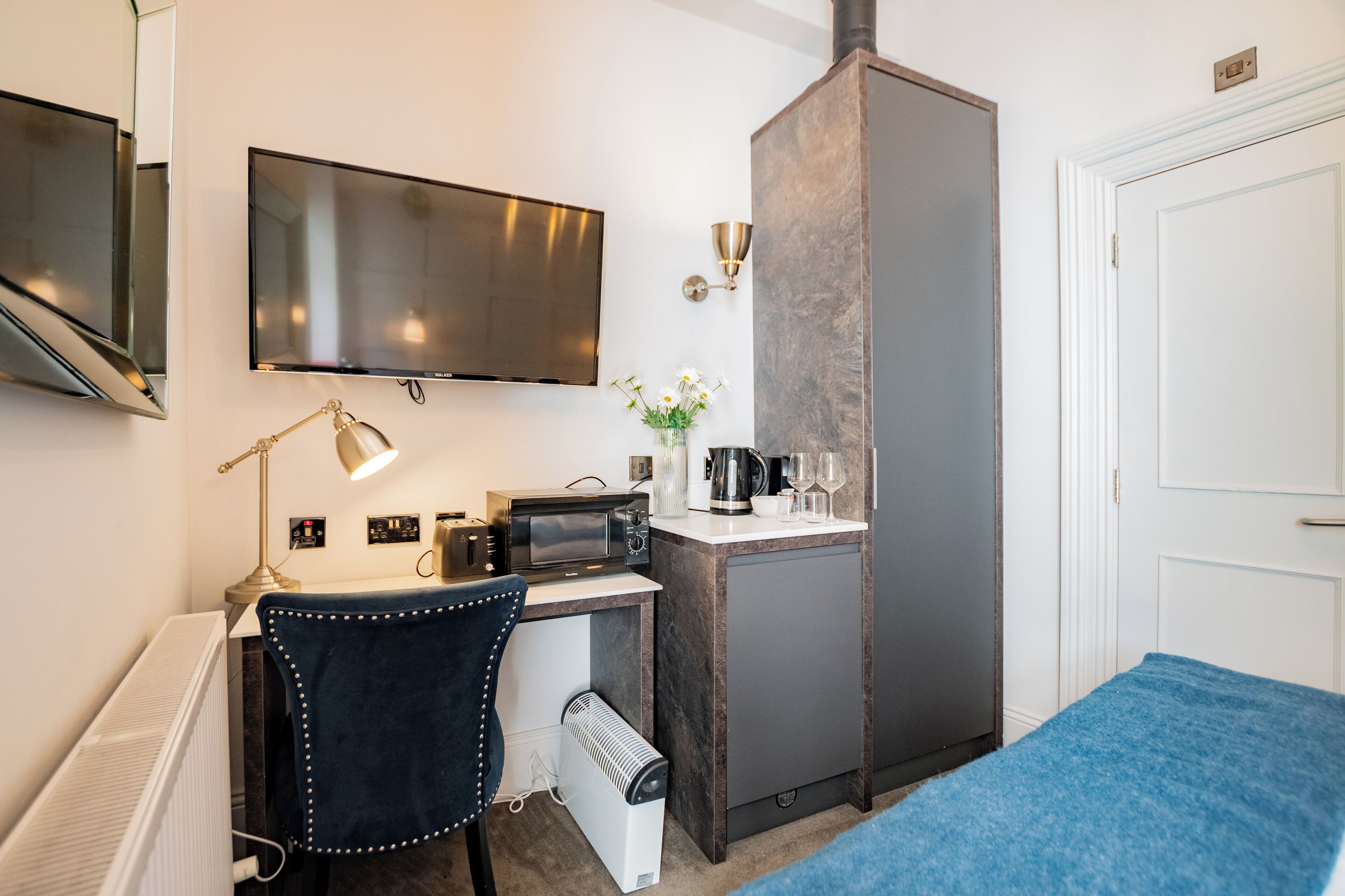 Kompakti hotellihuone, jossa on pieni keittiö, taulutelevisio, työpöytä lampun kanssa ja sänky, jossa on sininen päällinen.