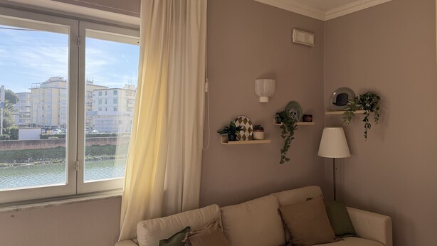 A living room with a sofa, a lamp, and a view of buildings through a window. - Hotel Laurin (Camaiore)