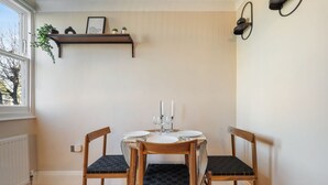 Ein Essbereich mit Tisch für zwei Personen, Holzstühle mit schwarzen Kissen, einem an der Wand befestigten Regal mit Pflanzen und einem Fenster mit Blick auf Bäume.
