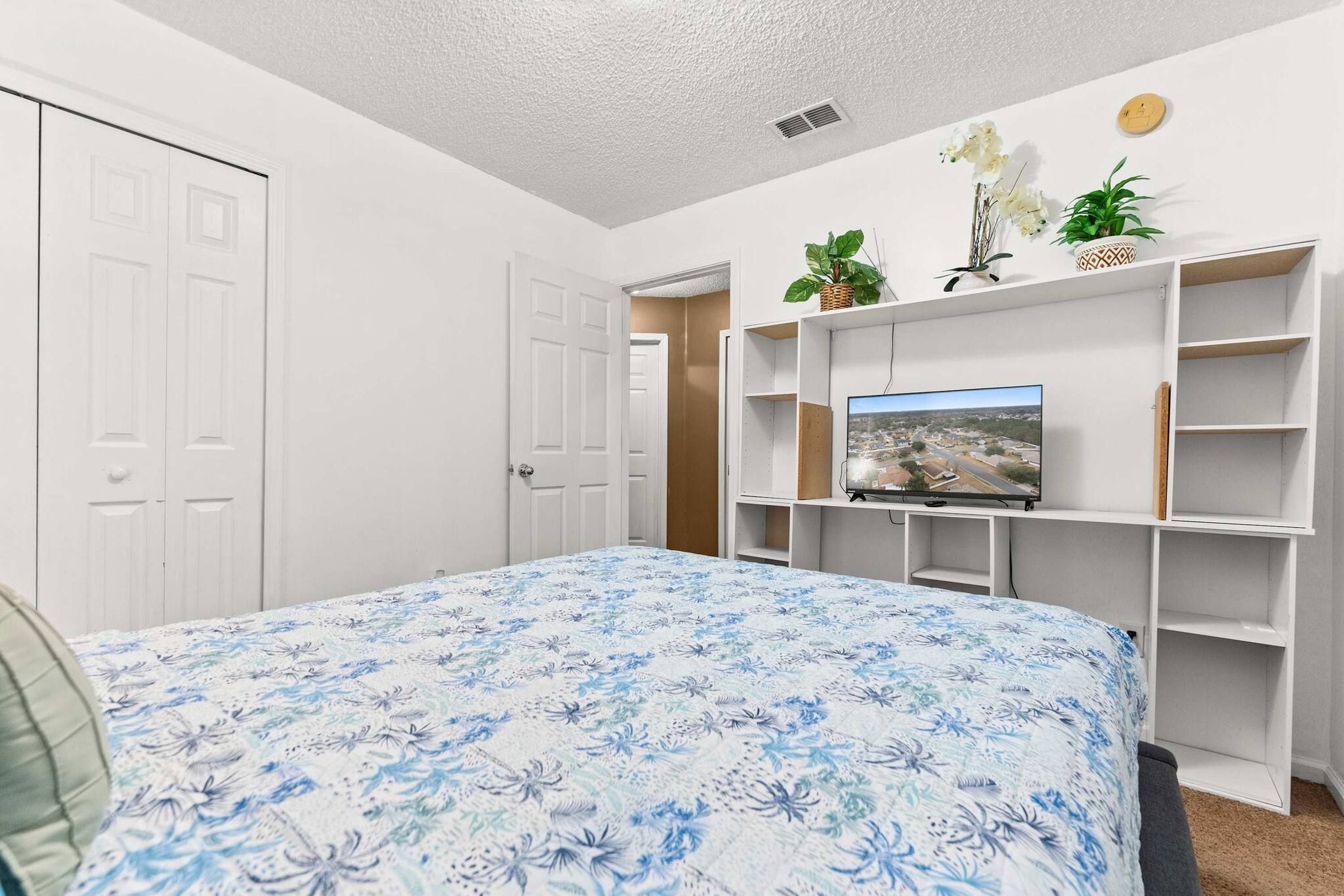 A bedroom with a bed, a television on a stand, a door, and a white wall with shelves.