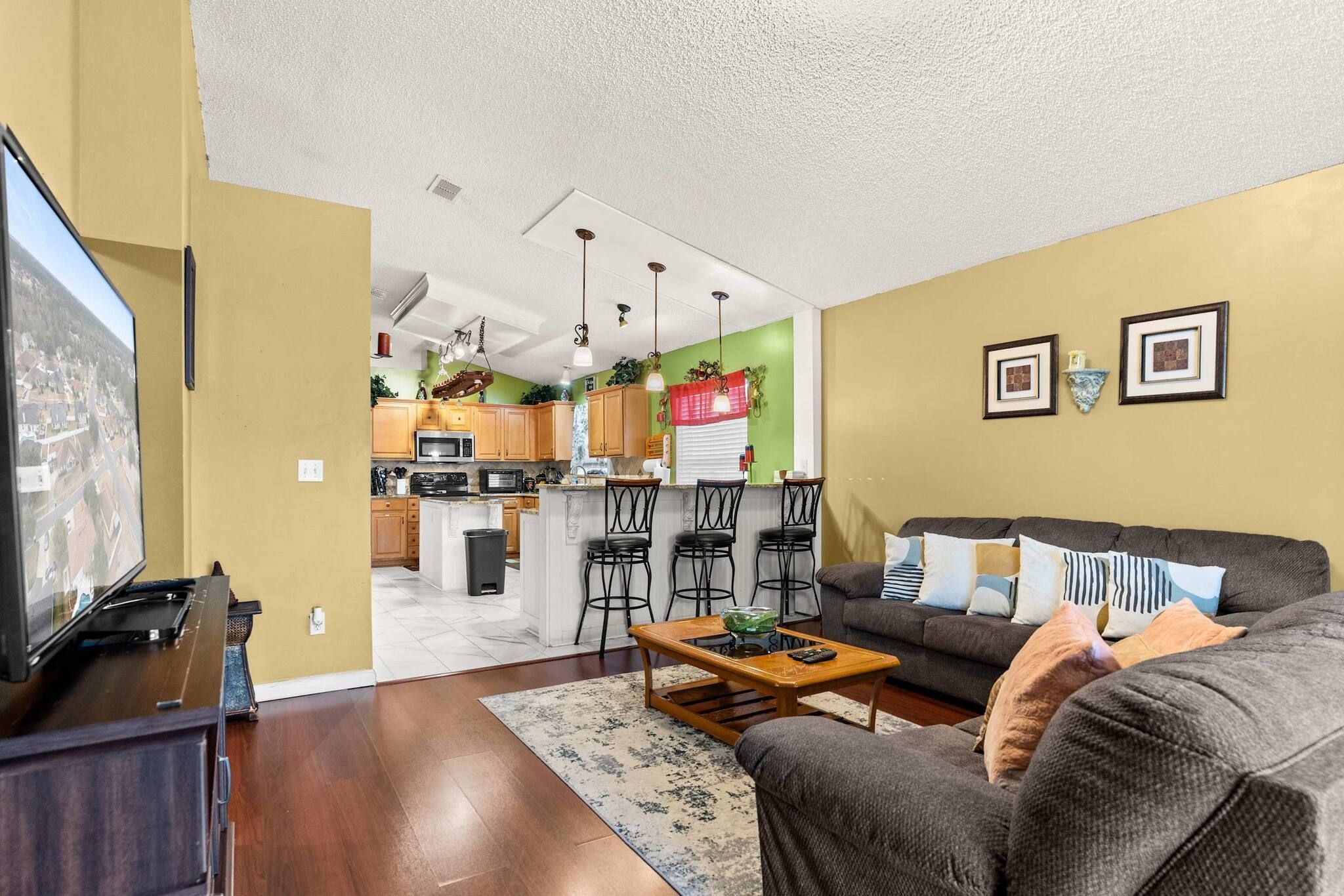 A living room with a sofa, a coffee table, and a TV. There is a kitchen in the background with a dining area.