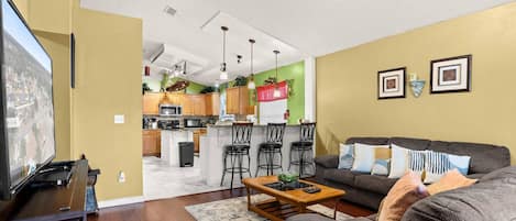 A living room with a sofa, a coffee table, and a TV. There is a kitchen in the background with a dining area.