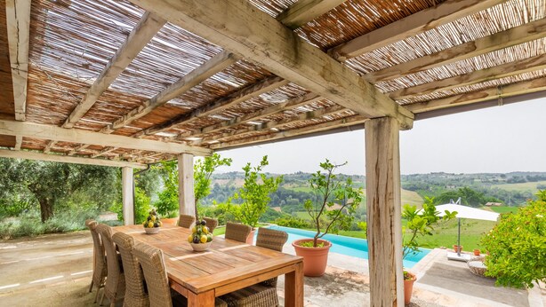 Eine überdachte Terrasse mit einem Holztisch, Korbstühlen, einem Topfpflanzen und Blick auf einen Pool und die Landschaft.