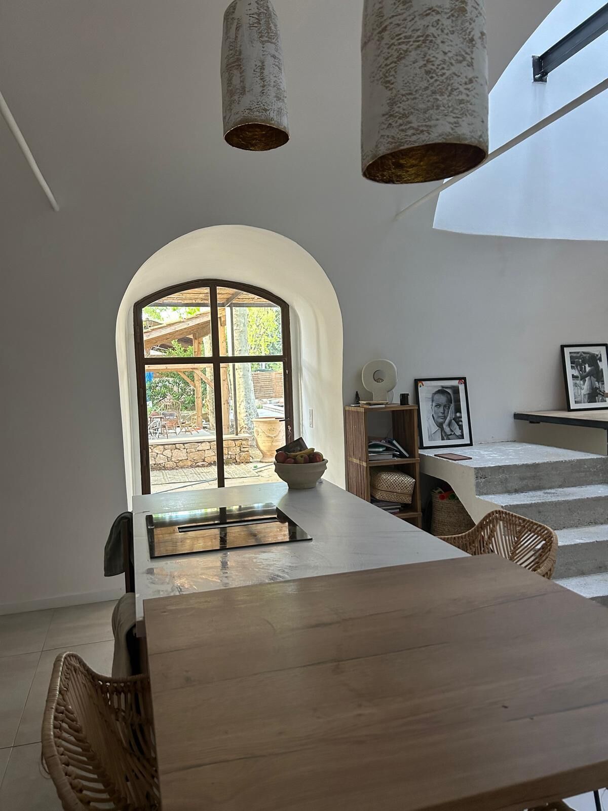 A dining area with a large table, wicker chairs, a bowl of fruit, and a view through an arched window to an outdoor patio.