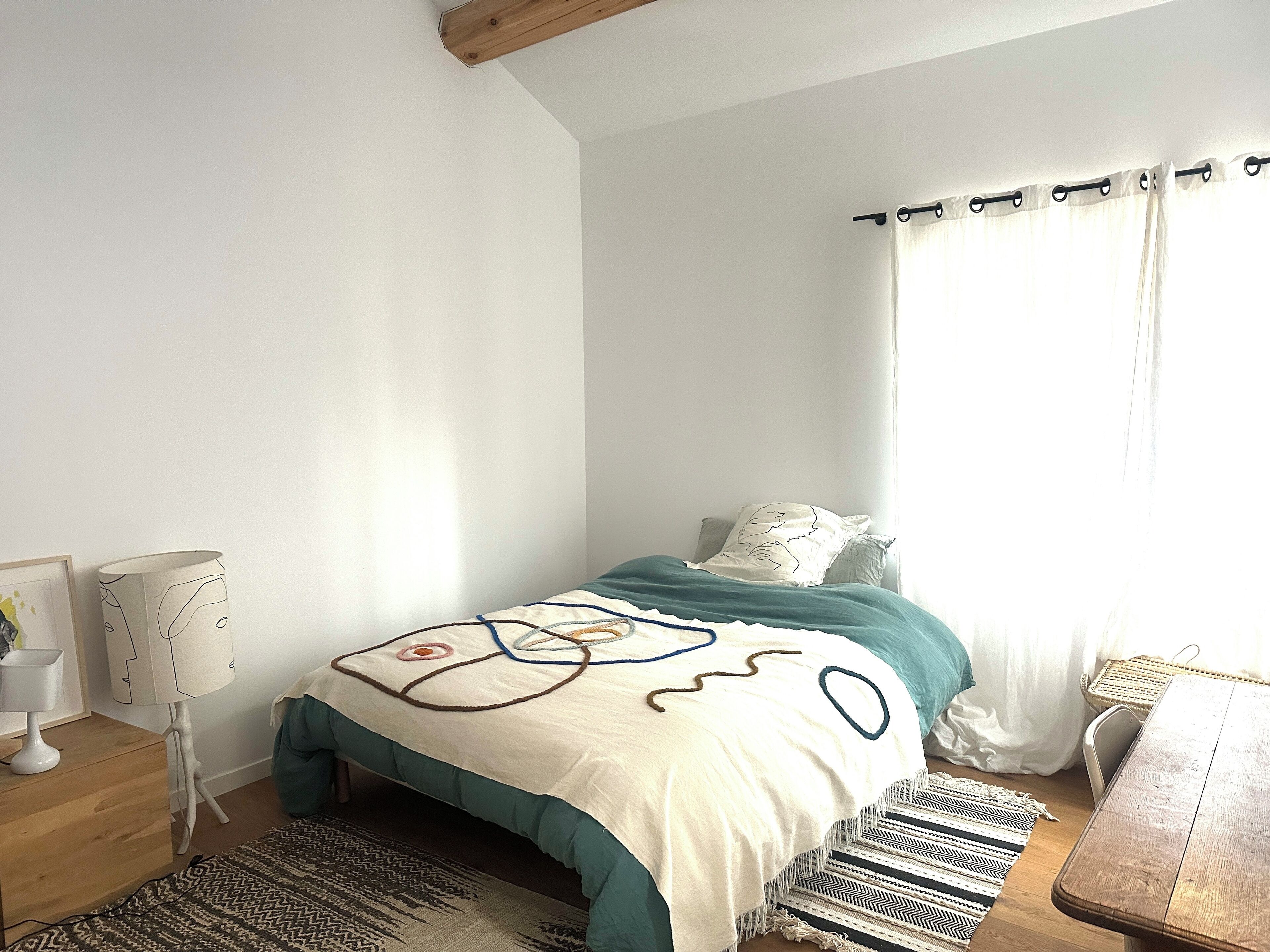 Een slaapkamer met een bed, nachtkastje, lamp, raam met gordijnen en een houten tafel.