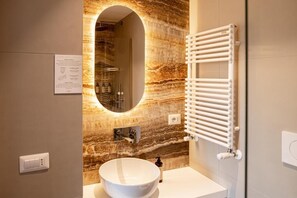 A modern bathroom with a marble backsplash, a round mirror, a white sink, and a towel rack. - Flora Guest House (Rome)