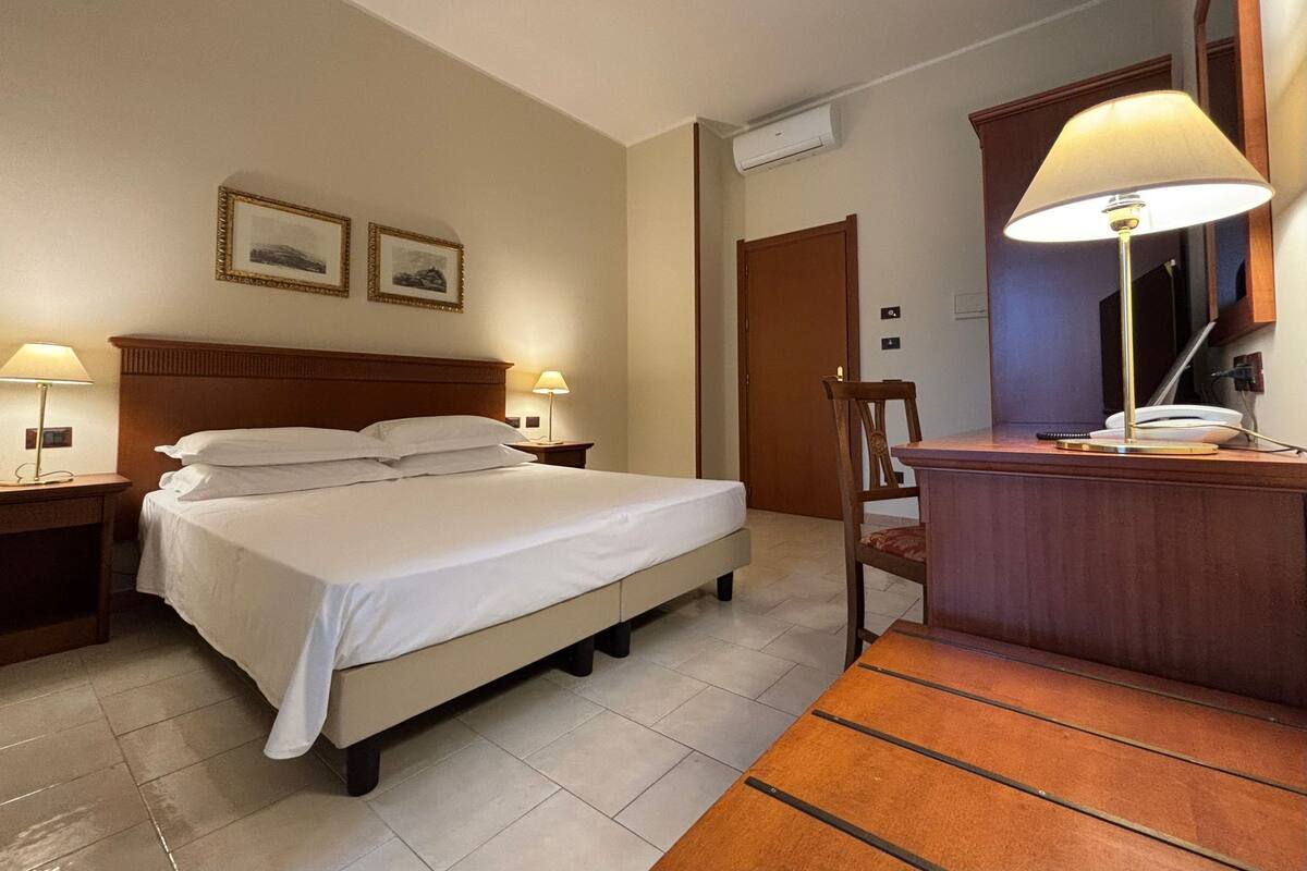 A hotel room with a bed, two bedside tables with lamps, a wooden desk with a lamp, and a chair.