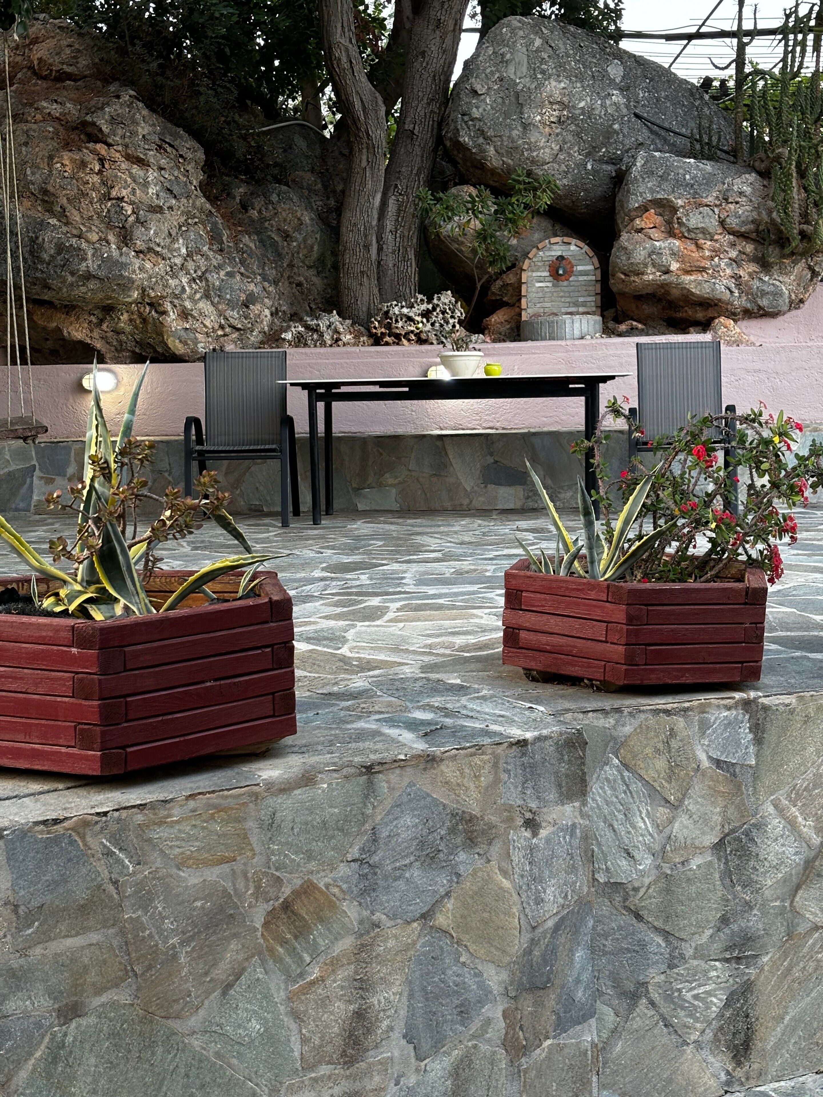 Ein Patio mit Steinboden, zwei hölzernen Pflanzkästen mit Pflanzen, einem Tisch und Stühlen.