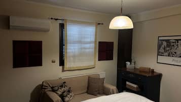 A hotel room with a bed, a window with a curtain, an air conditioning unit, a wall-mounted heater, a dark-colored dresser, and a framed artwork on the wall.
