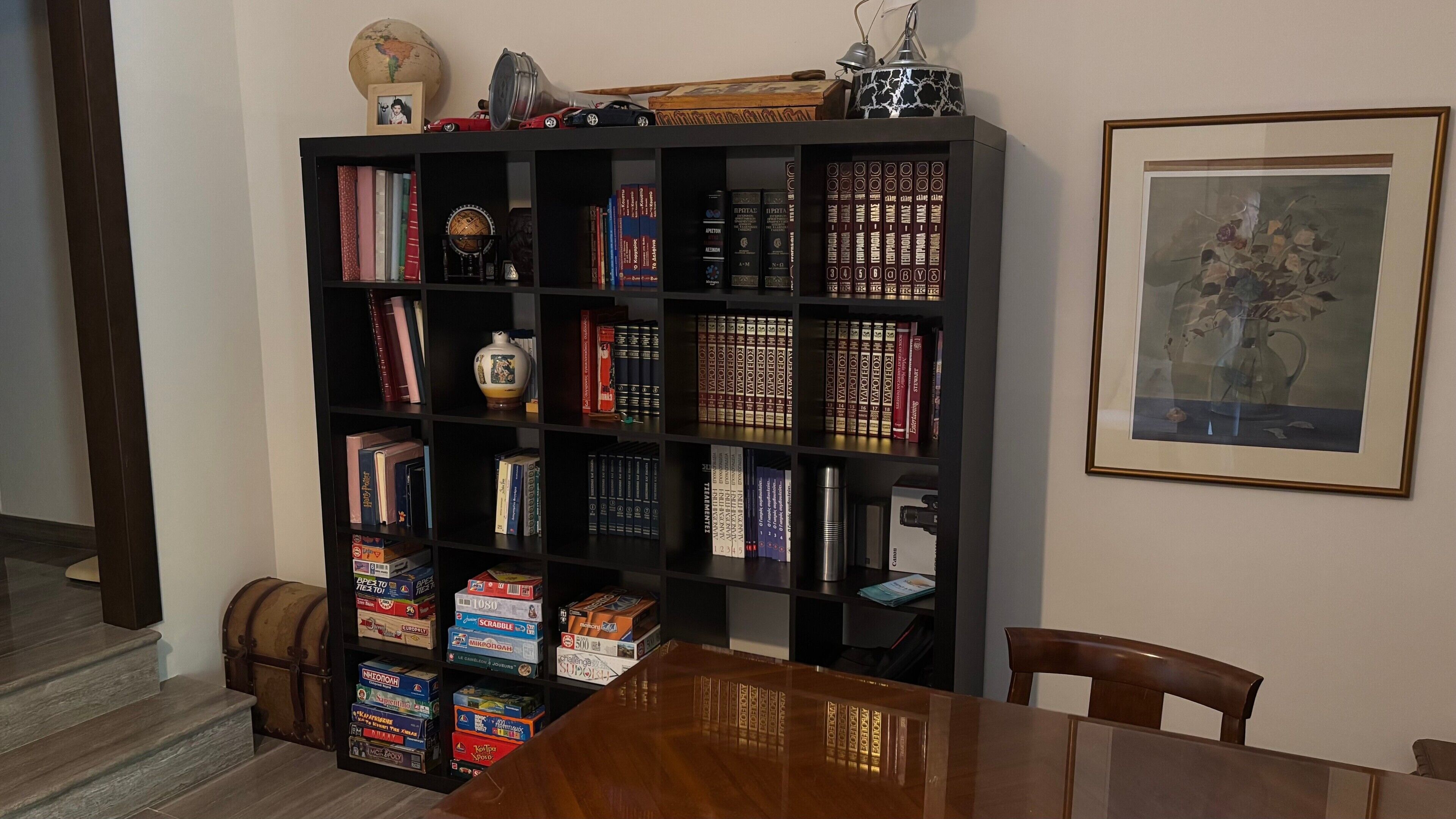 Une pièce avec une table en bois, une bibliothèque remplie de livres et de jeux de société, un globe terrestre et un tableau encadré accroché au mur.