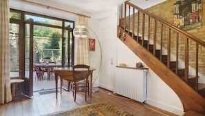 Un espace repas avec une table et des chaises en bois, un tapis à motifs, un escalier avec des rampes en bois et, à travers des portes vitrées, une vue sur un jardin extérieur.