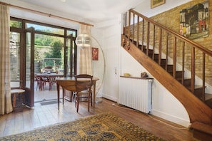 A dining area with a wooden table and chairs, a patterned rug, a staircase with wooden railings, and a view through glass doors to an outdoor garden. - Maison des papillons Daglan – Spacious Family Home with Pool in Village Center (Daglan)