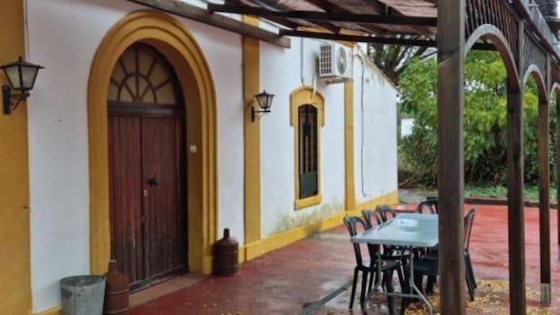 A covered outdoor area with a table and chairs, a wooden door, and a yellow and white building with arched windows.