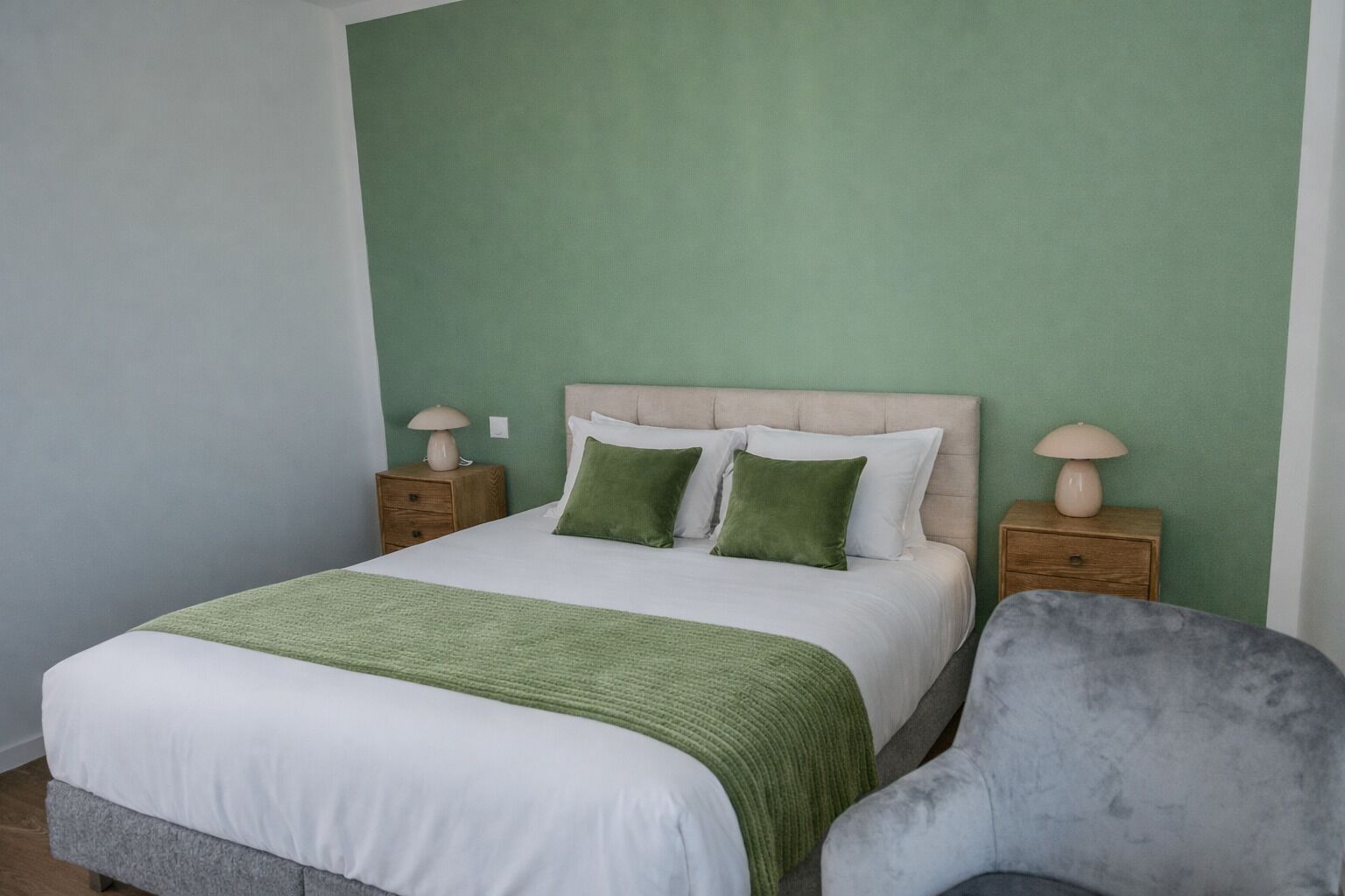 A neatly made bed with a green throw, two pillows, and a grey armchair in a room with green walls and wooden nightstands.