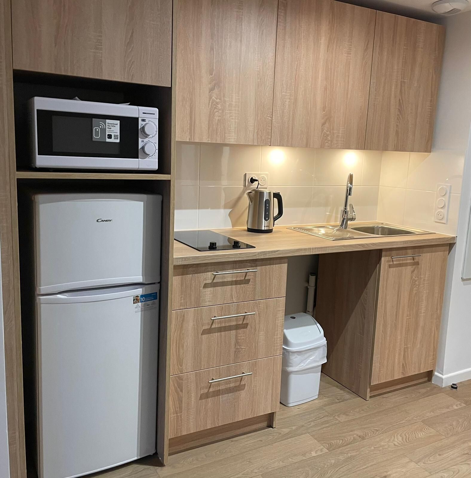 A compact kitchen with a microwave, refrigerator, kettle, sink, and wooden cabinets.